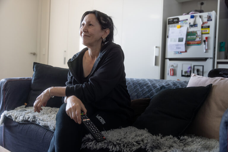 An older woman sits cross-legged on a lounge, smiling towards someone out of view.