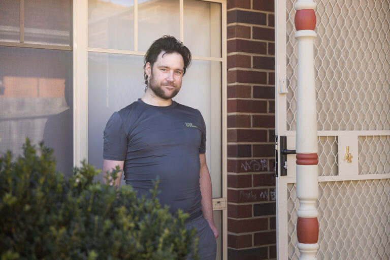 Young man with dark hair and a beard, standing in front of a door, smiling gently toward the camera.