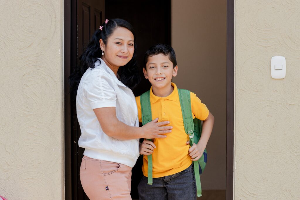 A child wearing a school uniform stands in a doorway, being hugged from the side by their mother, both looking towards the camera.